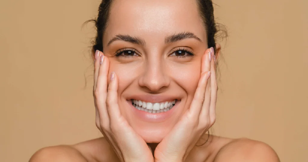 _Smiling woman with glowing, clear skin after MOXI in Murrysville, PA, gently holding her face with both hands against a neutral background.