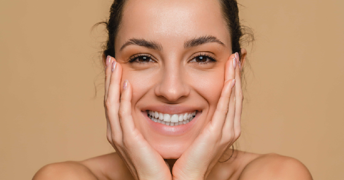 _Smiling woman with glowing, clear skin after MOXI in Murrysville, PA, gently holding her face with both hands against a neutral background.